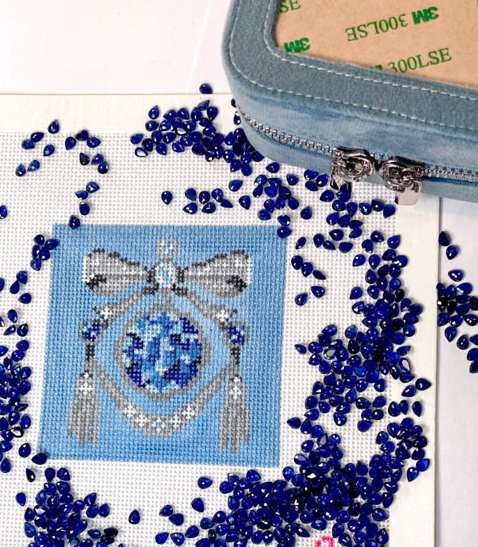 Blue beaded bow with central blue teardrop gemstone, on light fabric backing